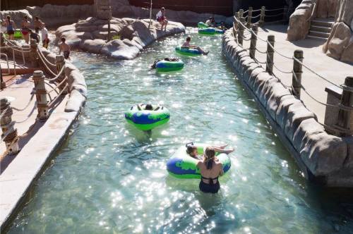 A lazy river with people floating on inner tubes, surrounded by rocky landscaping and sunlight reflecting on the water.