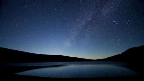 A serene night sky filled with stars reflects on a calm lake, surrounded by dark silhouettes of mountains.