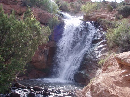 A cascading waterfall flows over rocky terrain, surrounded by lush greenery and red rock formations.