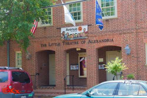 Exterior of the Little Theatre of Alexandria, featuring a sign for Avenue Q and flags outside a brick building.