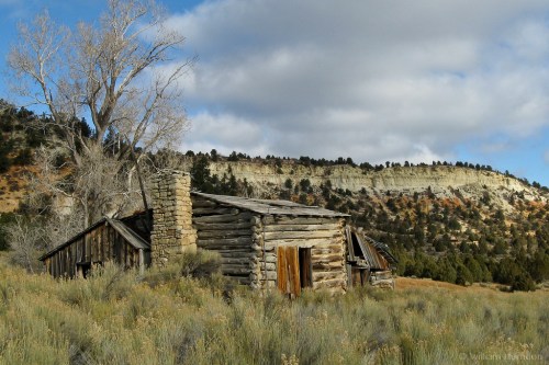 Abandoned log cabin in a grassy landscape, surrounded by hills and a cloudy sky.