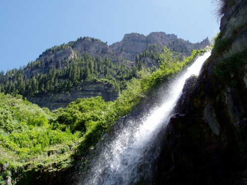 A waterfall cascades down a rocky cliff surrounded by lush greenery and towering mountains under a clear blue sky.