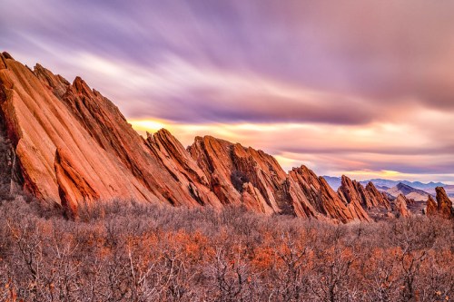 A dramatic landscape of jagged red rock formations under a colorful sky at sunset, surrounded by sparse vegetation.