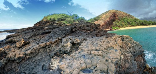 A panoramic view of rocky coastline with lush green hills and a calm blue sea under a partly cloudy sky.