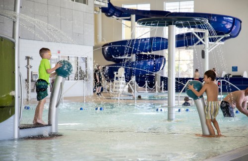 Two children play in a splash pad area with water features and slides in a bright indoor pool.