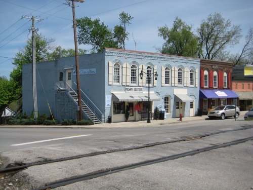 A quaint street scene featuring a blue building with shops and a car parked nearby, surrounded by greenery.
