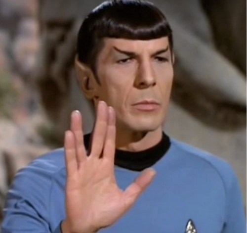 A character in a blue uniform raises a hand in a Vulcan salute, with pointed ears and a serious expression.