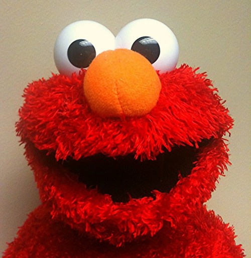 A close-up of a red, furry puppet with large eyes and an orange nose, smiling against a plain background.