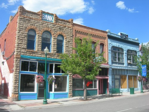 Historic buildings line a street, featuring a stone bank and colorful storefronts under a blue sky with clouds.