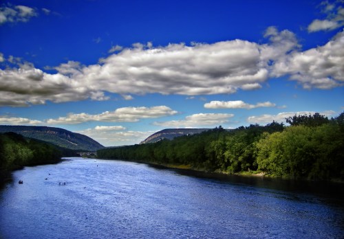 A serene river flows between lush green hills under a bright blue sky with fluffy clouds.