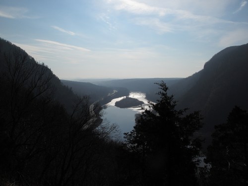 A serene river winding through a valley, surrounded by trees and mountains under a cloudy sky.