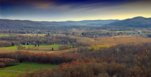 A panoramic view of rolling hills and valleys, with patches of green and autumn foliage under a colorful sky.