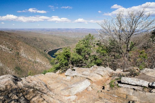 Scenic view from a rocky overlook, featuring a winding river and rolling hills under a blue sky with scattered clouds.