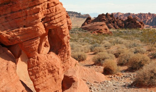A close-up of a red rock formation with desert vegetation and distant rocky hills under a clear sky.