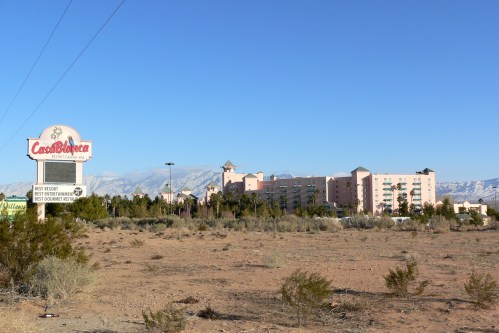 A desert landscape featuring a hotel with mountains in the background and a sign in front. Clear blue sky above.