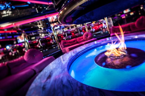 A stylish lounge with pink seating, vibrant lighting, and a circular fire pit surrounded by blue water.