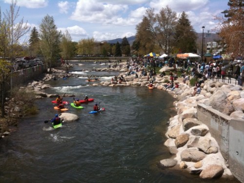 A river scene with kayakers navigating the water, surrounded by trees and a crowd enjoying a festival along the banks.