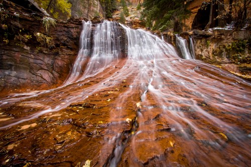 A cascading waterfall flows over rocky terrain, surrounded by autumn foliage and lush greenery.