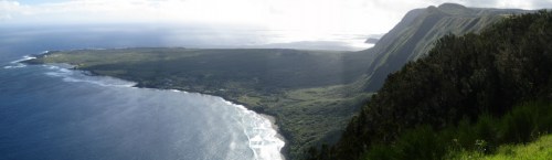 A panoramic view of a lush green coastline meeting the ocean, with dramatic cliffs and a cloudy sky in the background.