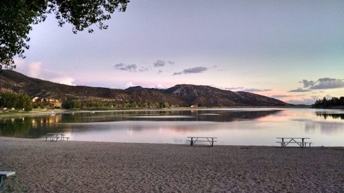 A serene lake at sunset, surrounded by mountains, with picnic tables on a sandy beach and a calm water reflection.