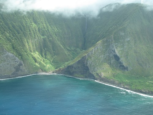 Lush green mountains rise above a blue ocean, with a waterfall cascading down the cliffs under a cloudy sky.