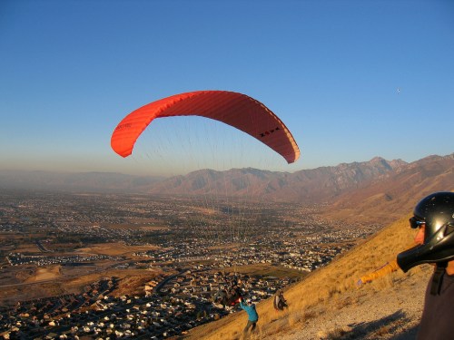 A paraglider with a red canopy soars above a valley, with mountains and a town visible below during sunset.