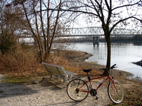 A red bicycle rests near a bench by a riverbank, with a bridge visible in the background and trees lining the shore.