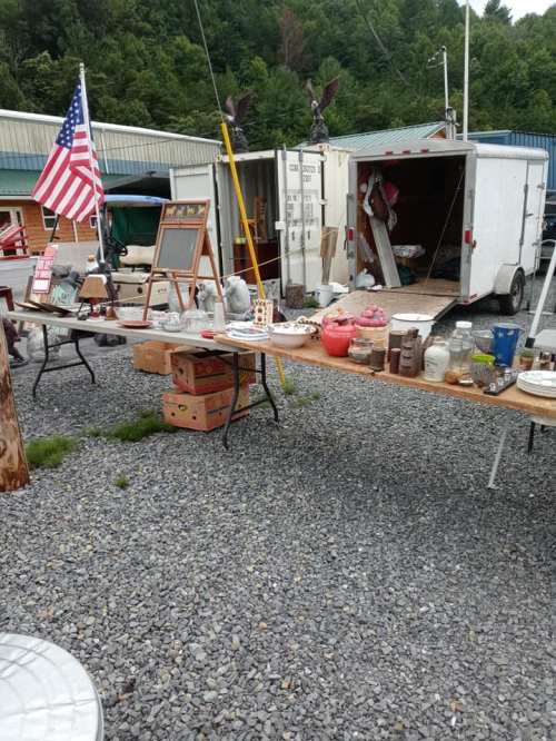 A bustling outdoor market scene with tables displaying various items, an American flag, and a trailer in the background.