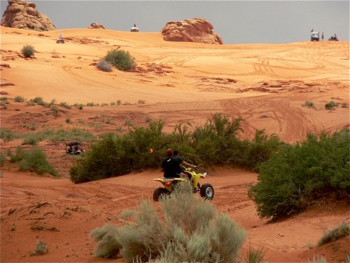 A person rides an ATV through sandy terrain with shrubs, while others are visible in the background on similar vehicles.
