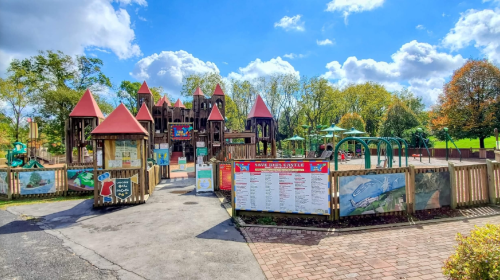Colorful playground with castle-like structures, surrounded by trees and a blue sky with fluffy clouds.