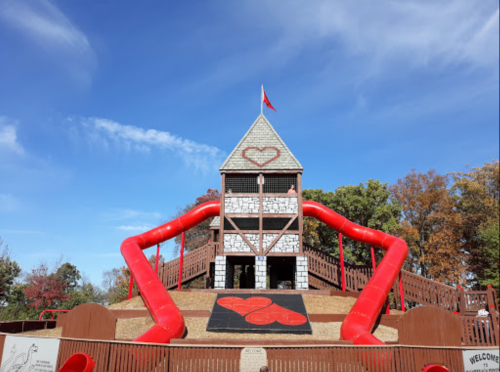 A playground structure resembling a castle with red slides and a heart design, surrounded by trees and blue sky.
