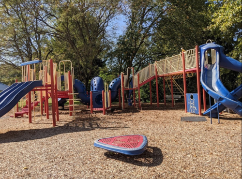 Colorful playground with slides, climbing structures, and a spinning disk, surrounded by trees and a sunny sky.