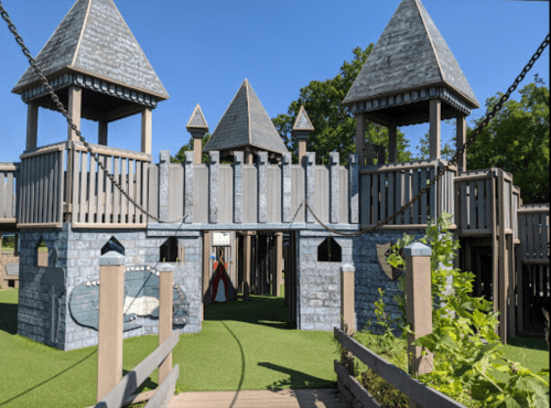 A large wooden playground resembling a castle, featuring towers, bridges, and green grass surrounding it.