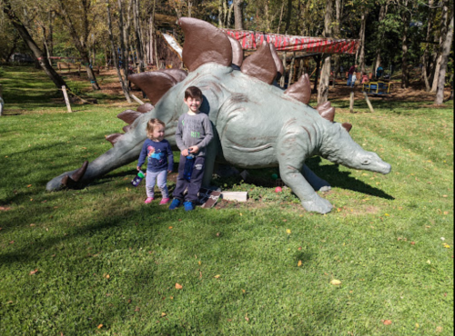 Two children pose next to a large, realistic dinosaur sculpture in a grassy park surrounded by trees.