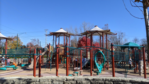 A colorful playground with various structures under a clear blue sky, surrounded by trees with autumn foliage.