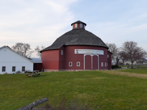 A round red barn with a thatched roof, surrounded by green grass and trees, under a cloudy sky.