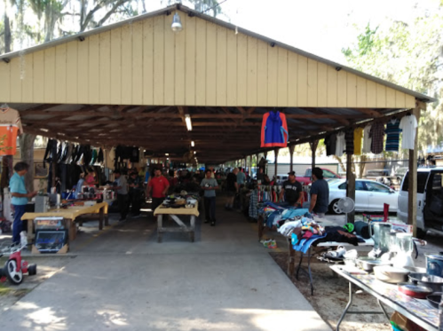 A bustling outdoor market with vendors selling clothes and various items under a large wooden shelter.