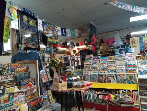 A cozy comic book store filled with shelves of comics, merchandise, and colorful banners hanging from the ceiling.