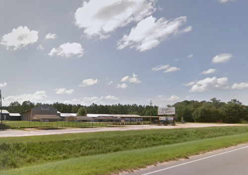 A roadside flea market with several buildings under a blue sky and fluffy clouds, surrounded by greenery.