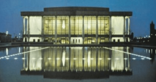 A modern building with large glass windows, reflecting in a calm water surface at dusk.