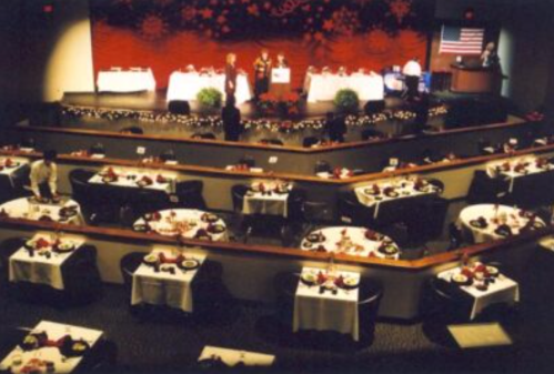 A banquet hall set for an event, featuring tables with white tablecloths and a stage with speakers.