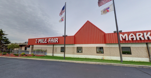 Exterior of 7 Mile Fair building with flags, featuring a red roof and large signage. Cloudy sky in the background.