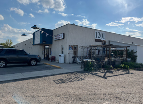 Exterior of a building with a sign, parked SUV, and outdoor seating area under a blue sky with clouds.