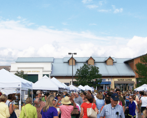 A bustling outdoor market with crowds of people and white tents under a blue sky with clouds.
