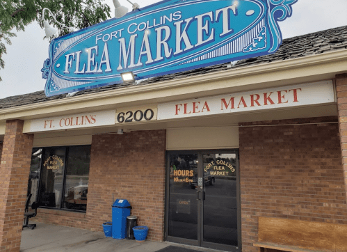 Sign for Fort Collins Flea Market, featuring blue lettering on a brick building with open hours displayed.