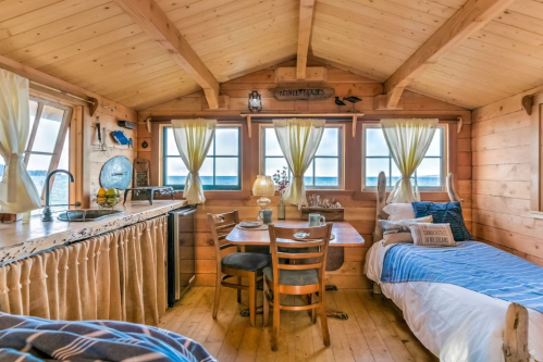 Cozy wooden cabin interior with a kitchen, dining table, and two beds, featuring large windows and a scenic view.
