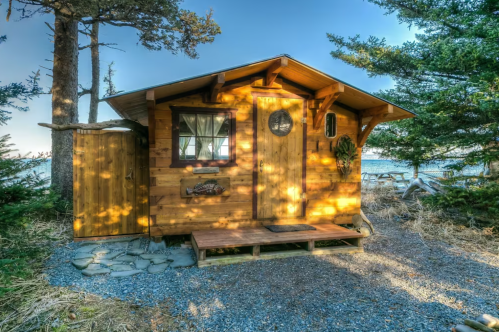 A cozy wooden cabin surrounded by trees, featuring a porch and a rustic door, set in a natural landscape.
