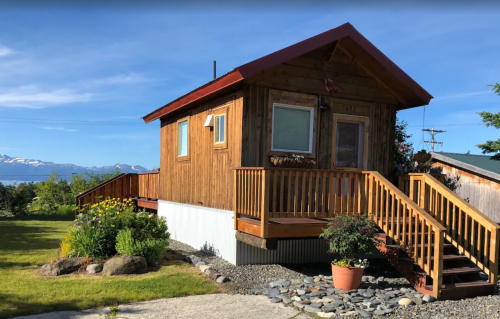 A cozy wooden cabin with a red roof, surrounded by greenery and flowers, overlooking a scenic landscape.
