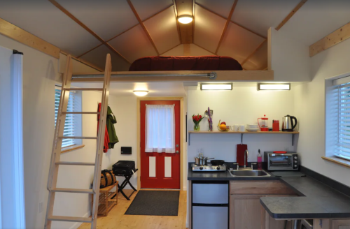 Cozy interior of a small cabin with a lofted bed, kitchen area, and a red door, featuring wooden accents and bright lighting.