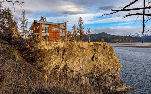 A modern wooden house perched on a rocky cliff by a calm body of water, surrounded by trees and mountains in the background.